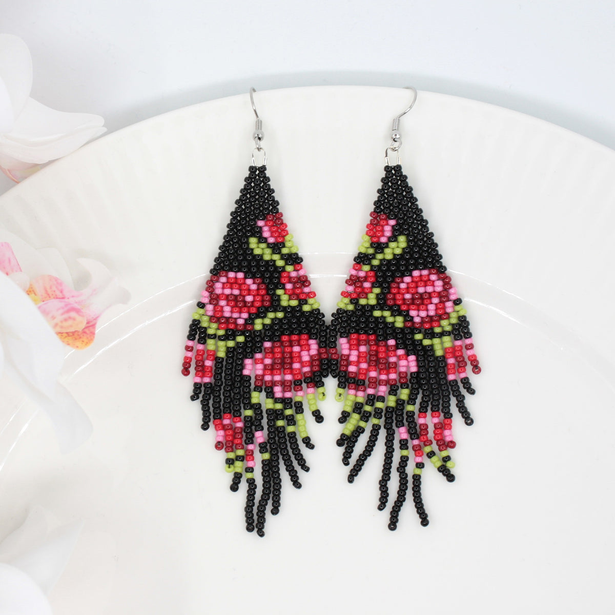 Long beaded seed bead fringe earrings on white plate. Base color of these earrings is and black and it has red roses pattern. Left side of the picture is white flowers. Pitkät käsintehdyt roikkuvat helmikorvakorut valkoisella lautasella. Pohjaväri näissä korvakoruissa on musta sekä, kuviona on punaisen sävyisiä ruusuja. Kuvan vasemmalla puolella on valkoisia kukkia.
