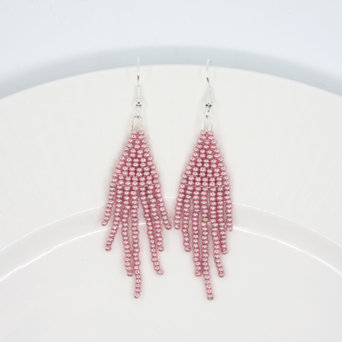 Hapsulliset helmikorvakorut värinä metallinen vaaleanpunainen valkoisella lautasella. Metallic pink-colored beaded seed bead fringe earrings on a white plate. 