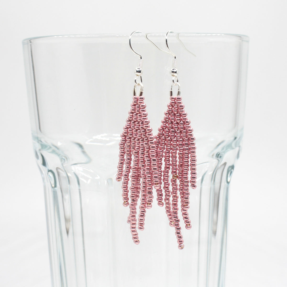 Hapsulliset helmikorvakorut värinä metallinen vaaleanpunainen roikkumassa juomalasin reunalla. Metallic pink-colored beaded seed bead fringe earrings hanging from the rim of a drinking glass.