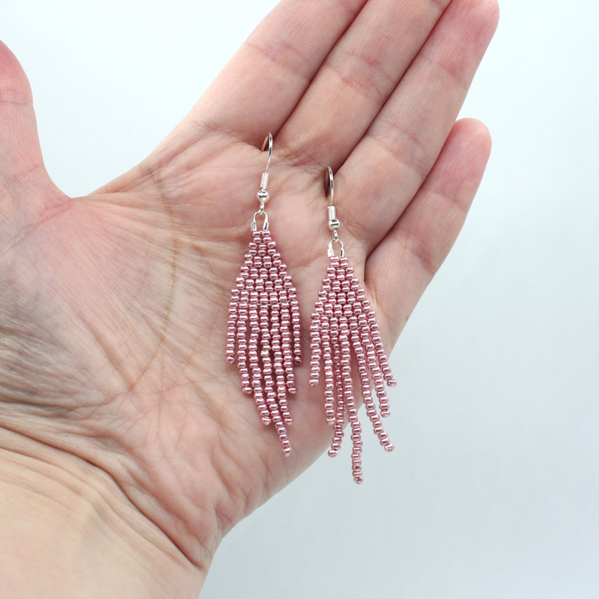 Hapsulliset helmikorvakorut värinä metallinen vaaleanpunainen roikkumassa kädessä. Metallic pink-colored beaded seed bead fringe earrings hanging from the hand.