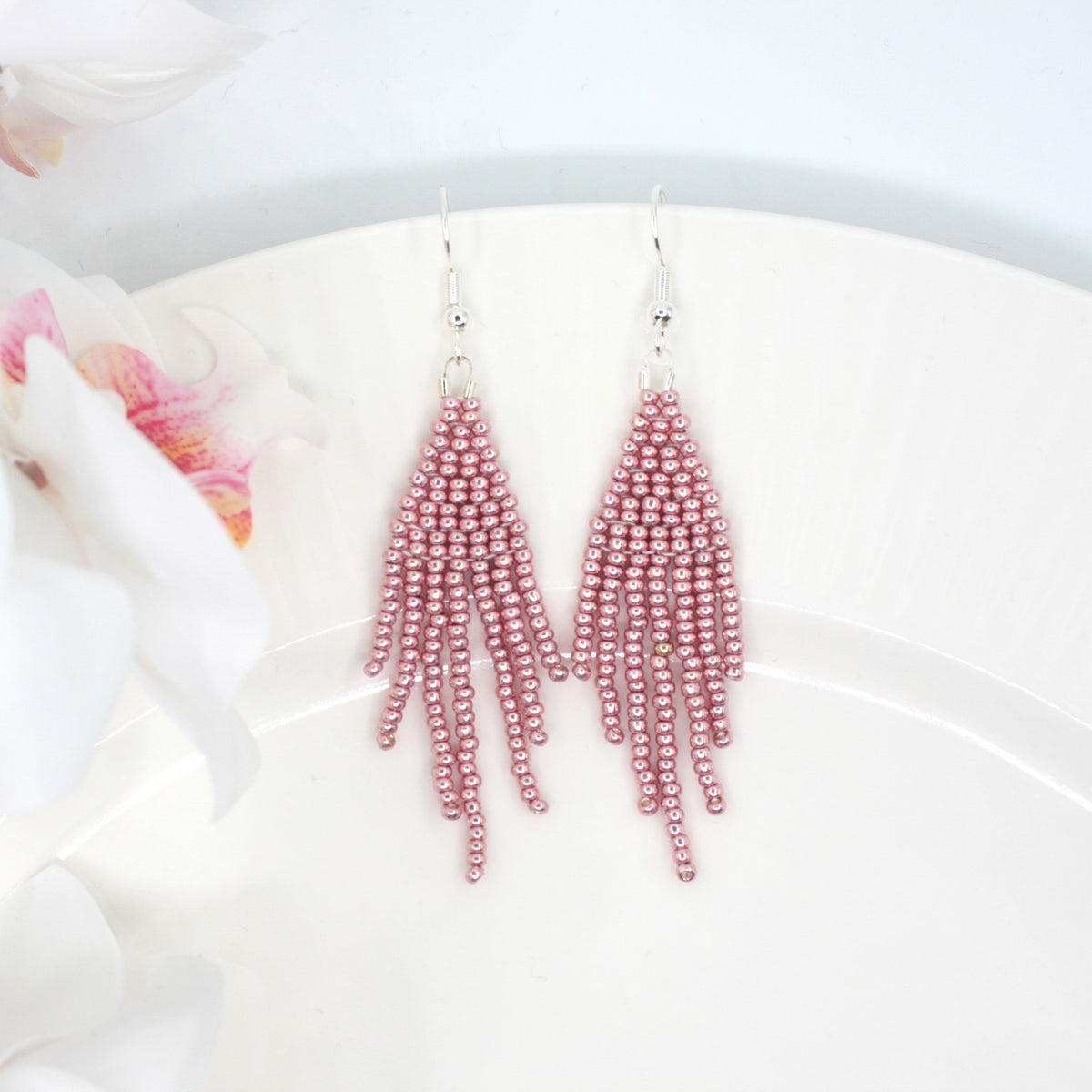 Hapsulliset helmikorvakorut värinä metallinen vaaleanpunainen valkoisella lautasella. Vasemmalla puolella näkyy hieman valkoisia kukkia. Metallic pink-colored beaded seed bead fringe earrings on a white plate. In the picture left side is little bit white flowers.