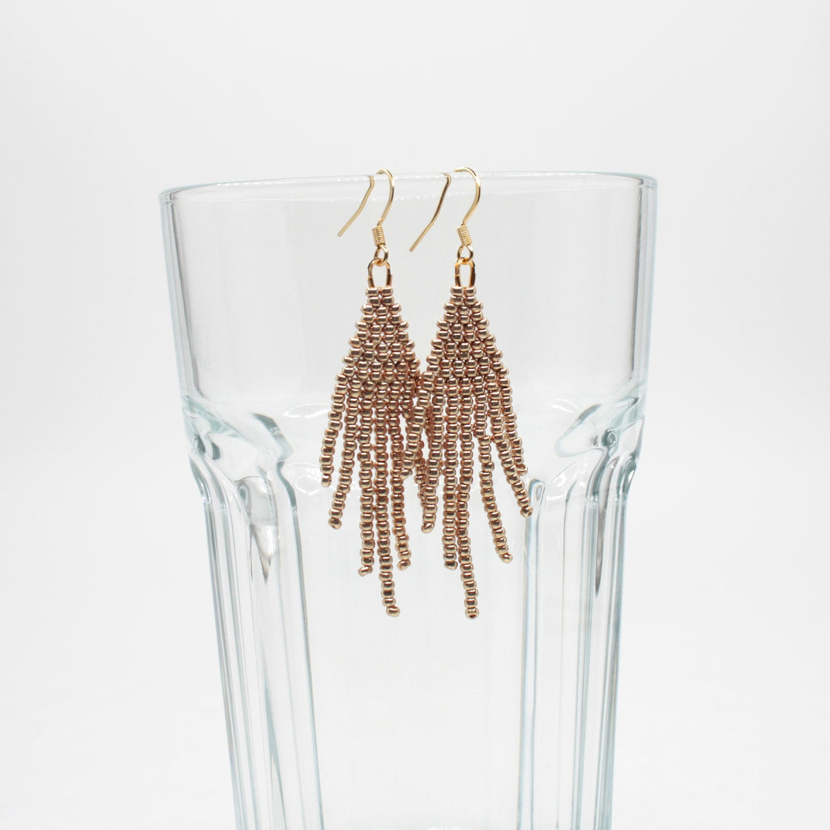 Hapsulliset helmikorvakorut värinä samppanja roikkumassa juomalasin reunalla. Champagne-colored beaded seed bead fringe earrings hanging from the rim of a drinking glass.