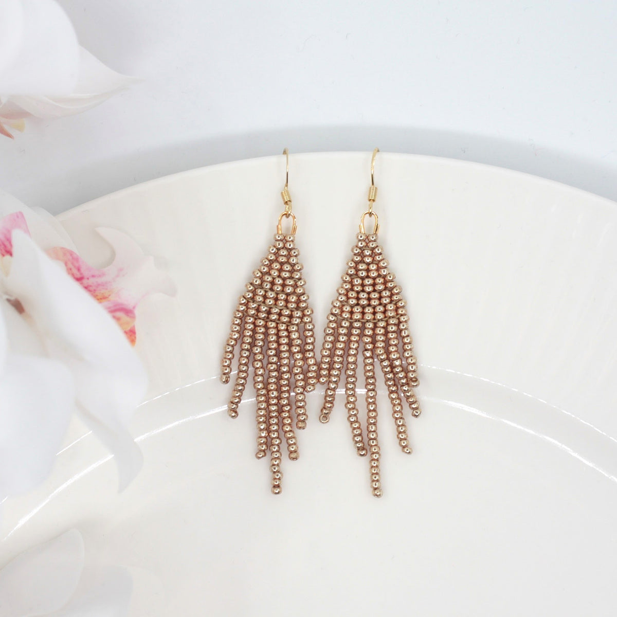Hapsulliset helmikorvakorut värinä samppanja valkoisella lautasella. Vasemmalla puolella näkyy valkoista kukkaa. Champagne-colored beaded seed bead fringe earrings on a white plate. A white flower is visible on the left side.