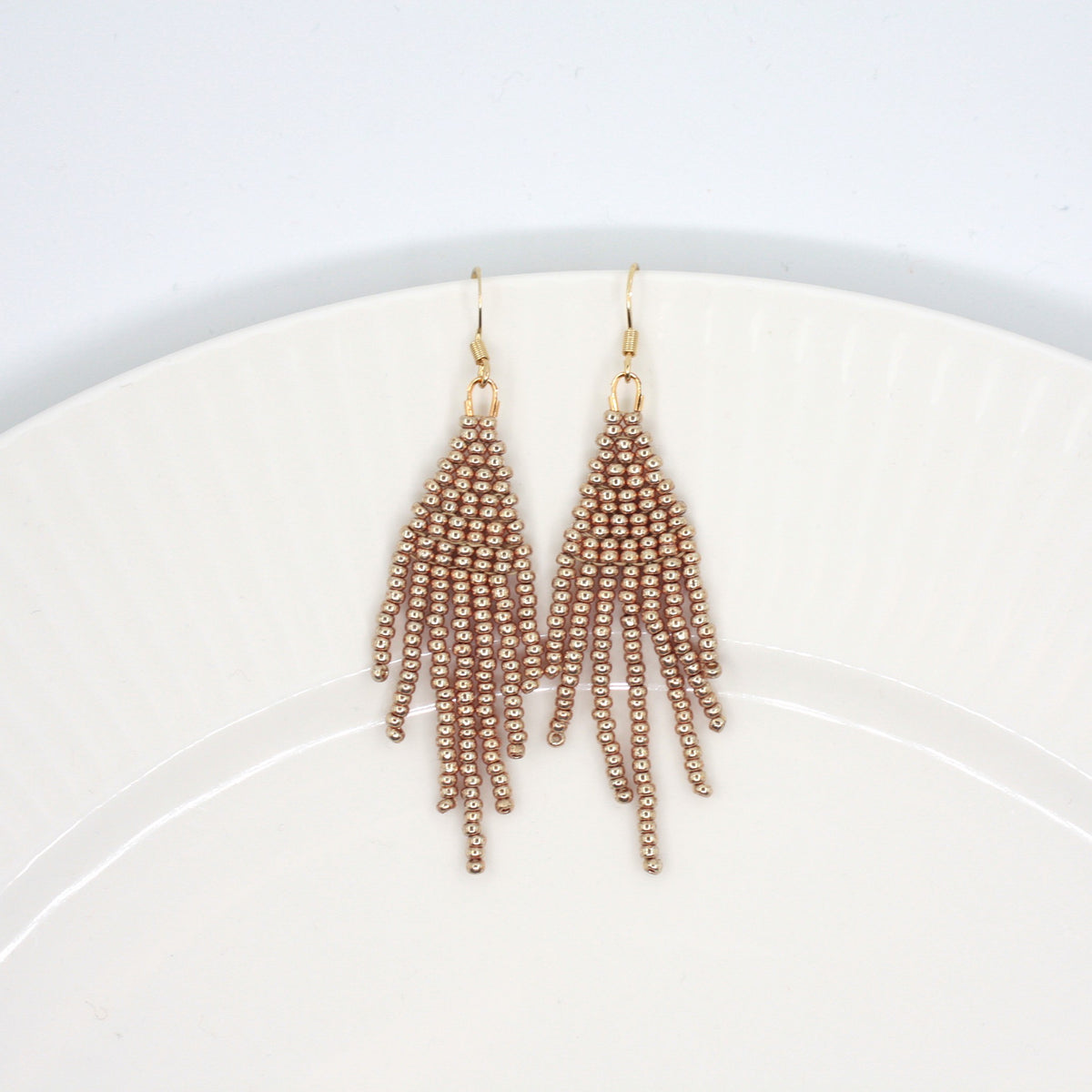 Hapsulliset helmikorvakorut värinä samppanja valkoisella lautasella. Champagne-colored beaded seed bead fringe earrings on a white plate.