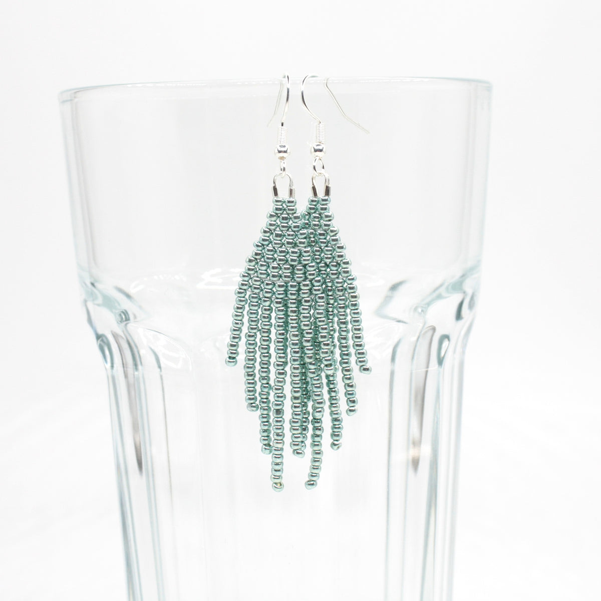 Hapsulliset helmikorvakorut värinä metallinen vaalean sininen roikkumassa juomalasin reunalla. Metallic light blue-colored beaded seed bead fringe earrings hanging from the rim of a drinking glass.