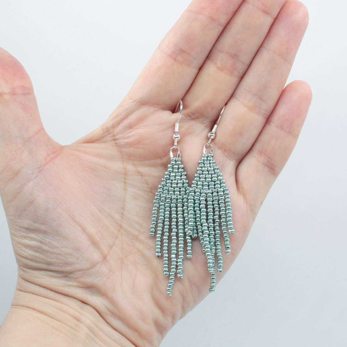 Hapsulliset helmikorvakorut värinä metallinen vaaleansininen roikkumassa kädessä. Metallic light blue-colored beaded seed bead fringe earrings hanging from the hand.
