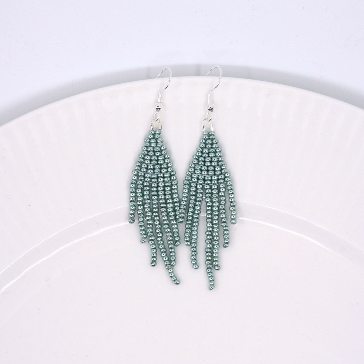 Hapsulliset helmikorvakorut värinä metallinen vaaleansininen valkoisella lautasella. Metallic light blue-colored beaded seed bead fringe earrings on a white plate. 