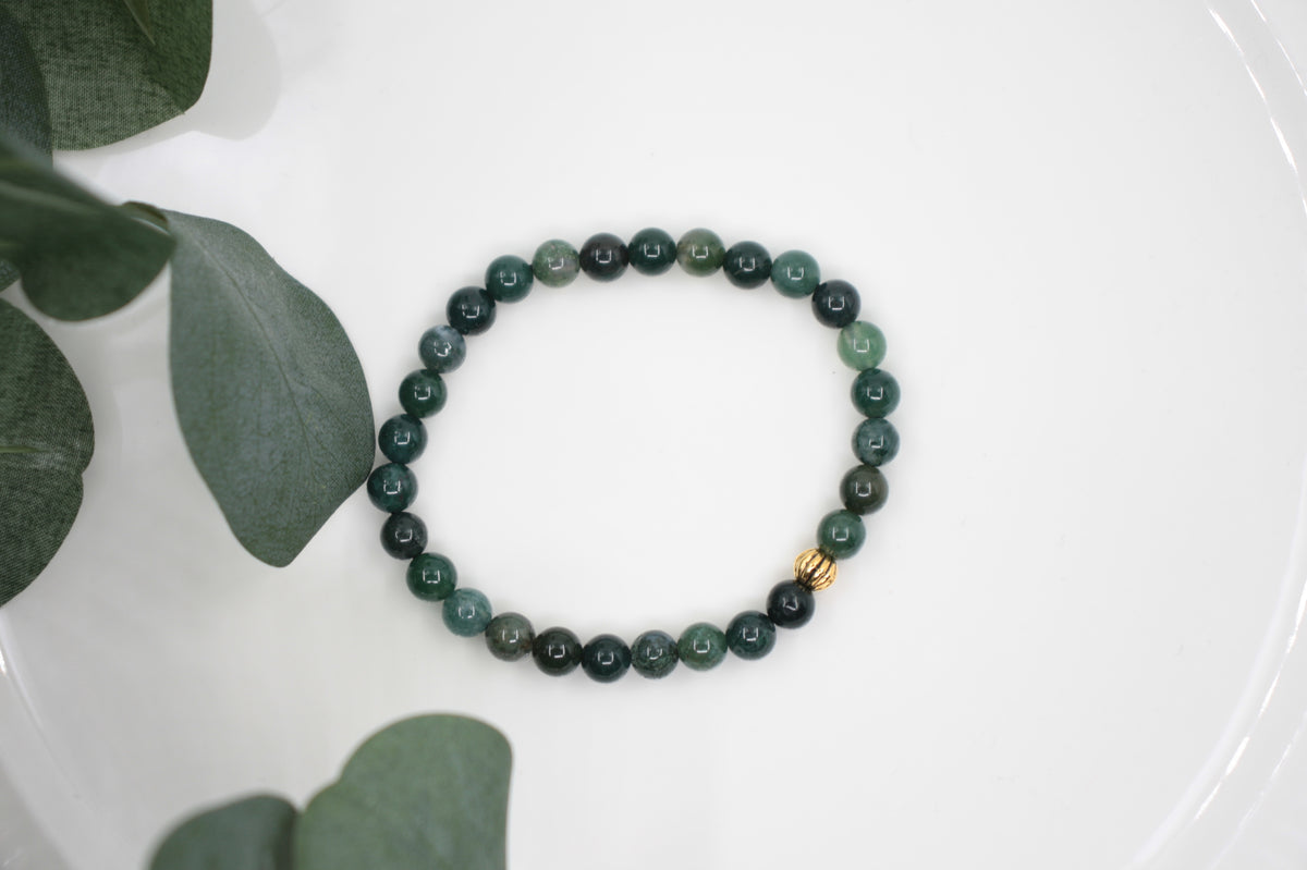 Vihreä sammalakaatti rannekoru kullanvärisellä metallihelmellä. Tausta on valkoinen ja sivulla vihreitä lehtiä. Green moss agate bracelet with gold color metal bead. Background is white and there is green leaves at the left side. 