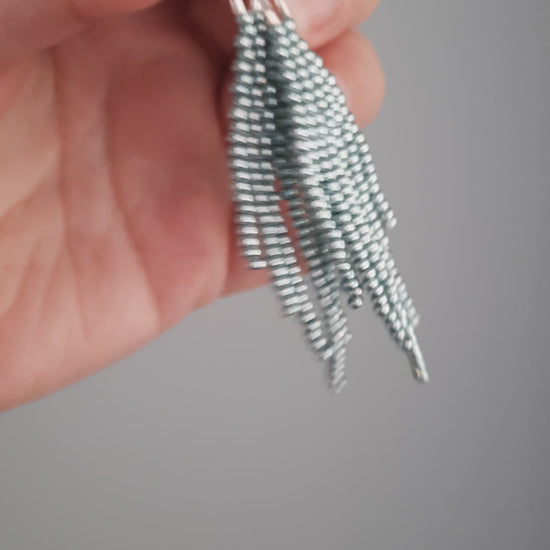 Hapsulliset helmikorvakorut värinä metallinen vaaleansininen roikkumassa kädessä. Metallic light blue-colored beaded seed bead fringe earrings hanging from the hand.