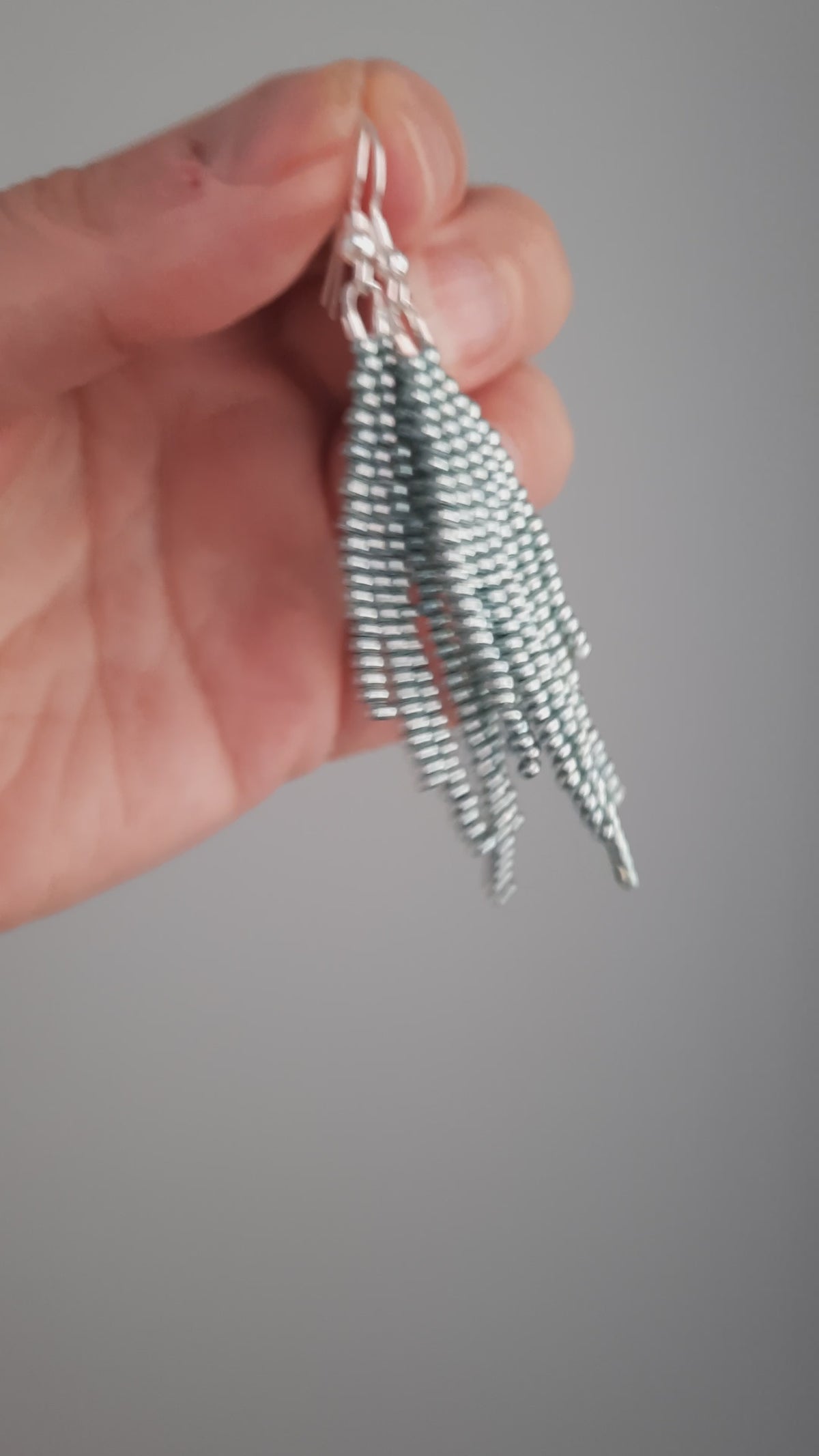 Hapsulliset helmikorvakorut värinä metallinen vaaleansininen roikkumassa kädessä. Metallic light blue-colored beaded seed bead fringe earrings hanging from the hand.