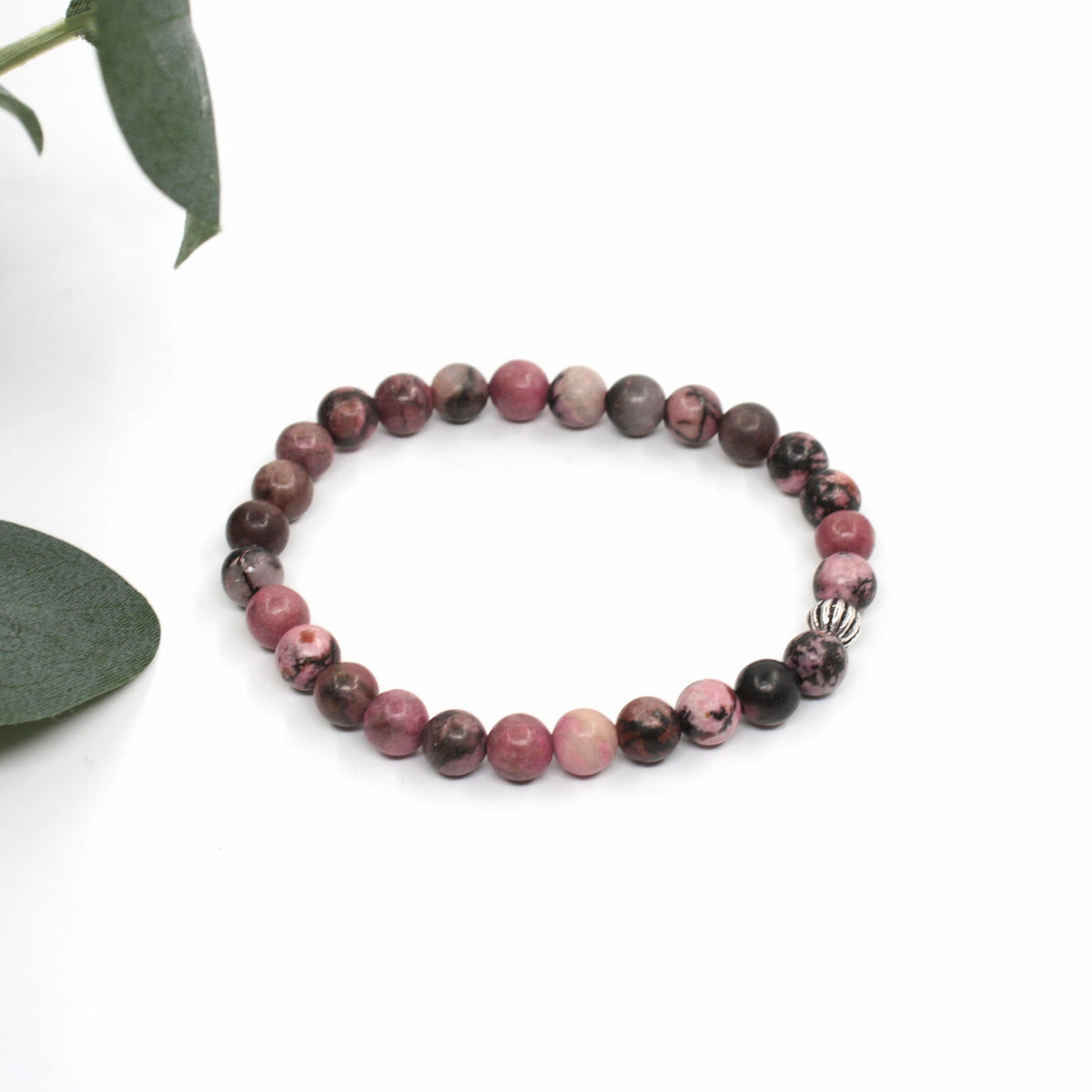 Rodoniitti rannekoru valkoisella alustalla. Vasemmalla puolella kuvassa näkyy vähän vihreitä lehtiä. Rhodonite bracelet on a white surface. A few green leaves are visible on the left side of the image.