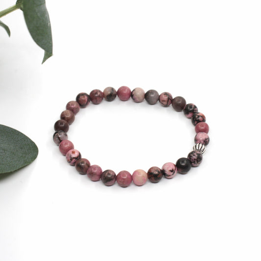 Rodoniitti rannekoru valkoisella alustalla. Vasemmalla puolella kuvassa näkyy vähän vihreitä lehtiä. Rhodonite bracelet on a white surface. A few green leaves are visible on the left side of the image.