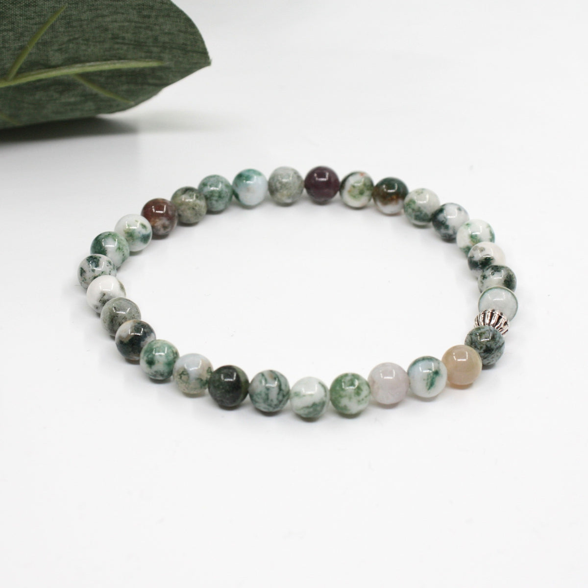 Puu-akaatti rannekoru valkoisella alustalla. Vasemmalla puolella kuvassa näkyy vähän vihreitä lehtiä. Wood agate bracelet on a white surface. A few green leaves are visible on the left side of the image.