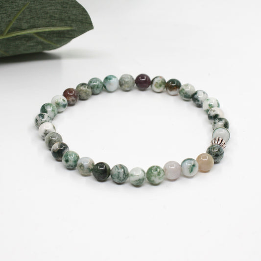 Puu-akaatti rannekoru valkoisella alustalla. Vasemmalla puolella kuvassa näkyy vähän vihreitä lehtiä. Wood agate bracelet on a white surface. A few green leaves are visible on the left side of the image.